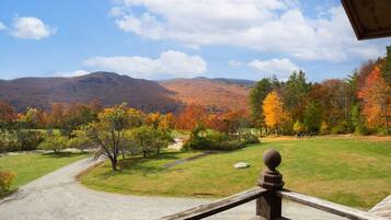 A scenic view from a balcony overlooking a grassy area with trees displaying autumn colors, a gravel path, and mountains in the background.