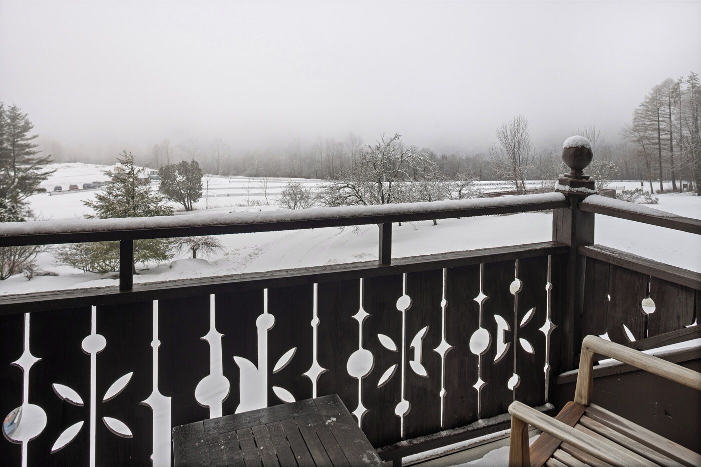 A wooden balcony with a decorative railing, overlooking a snowy landscape with trees and a distant view of a house.