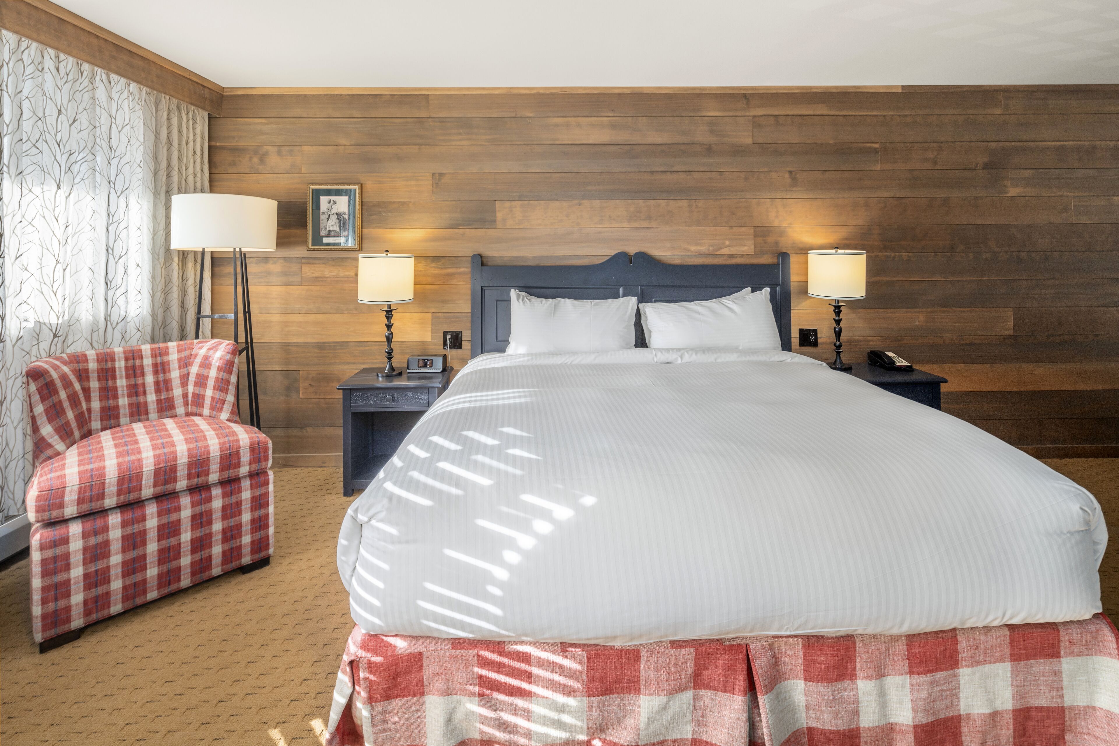 A hotel room with a large bed, two bedside tables, a red plaid armchair, and a wooden headboard.