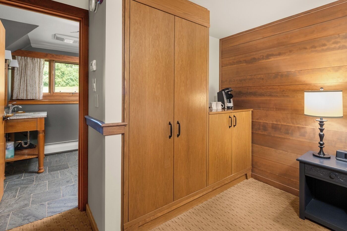 Een houten garderobe met twee deuren, een tafeltje met een lamp en een raam met gordijnen.