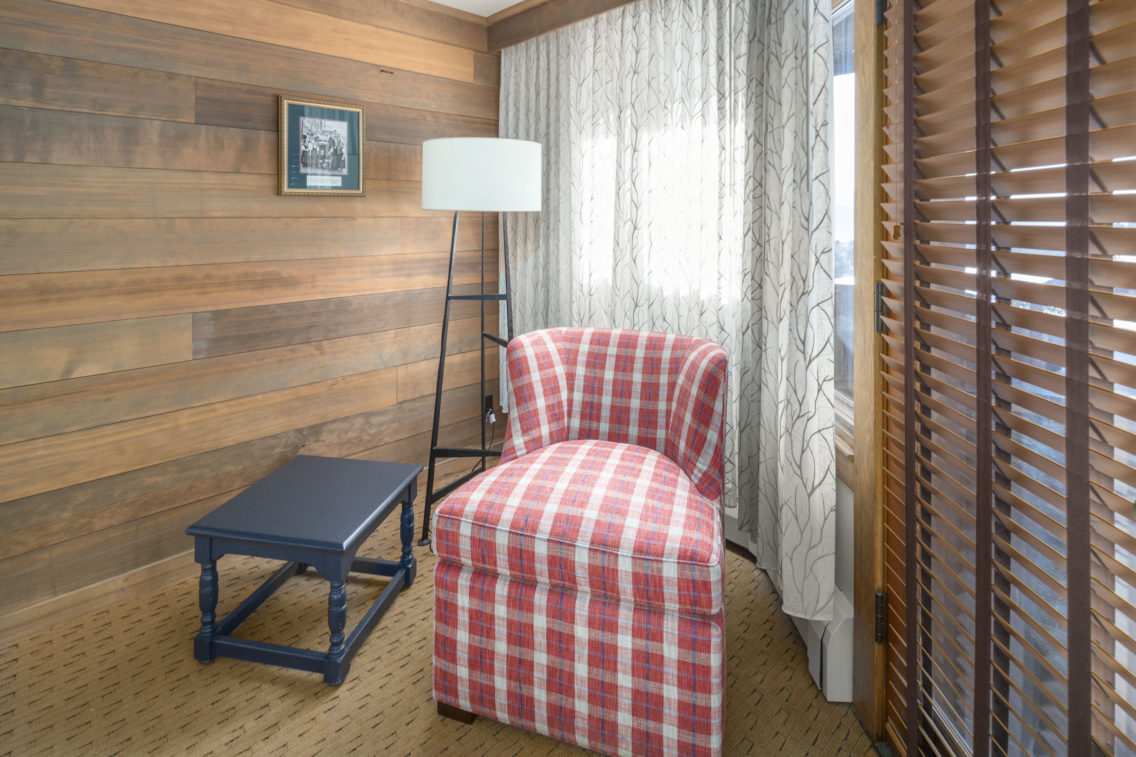 A room with a plaid chair, a small table, a lamp, and a window with blinds.