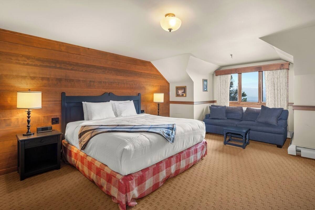 Una habitación de hotel con una cama grande, un sofá, una mesita de noche, una lámpara y una ventana con cortinas.