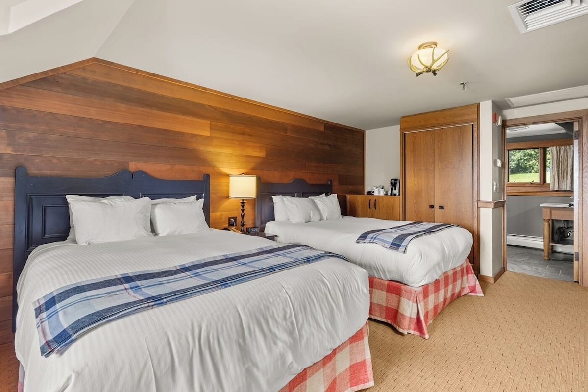 Habitación de hotel con dos camas, cabecero de madera, mesita de noche con lámpara, armario y vista al exterior a través de una ventana.