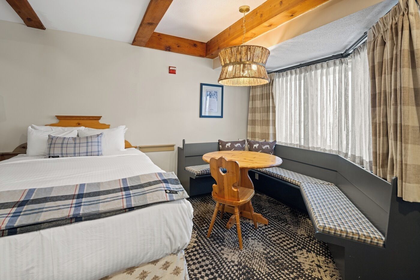 Ein Hotelzimmer mit einem Bett, einem runden Holztisch mit Stühlen, einer Bank mit Kissen und einem Fenster mit Vorhängen.