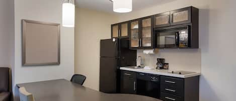 A modern kitchen with dark cabinetry, a stainless steel refrigerator, a microwave, and a coffee maker. There is a dining table with chairs and pendant lights above.