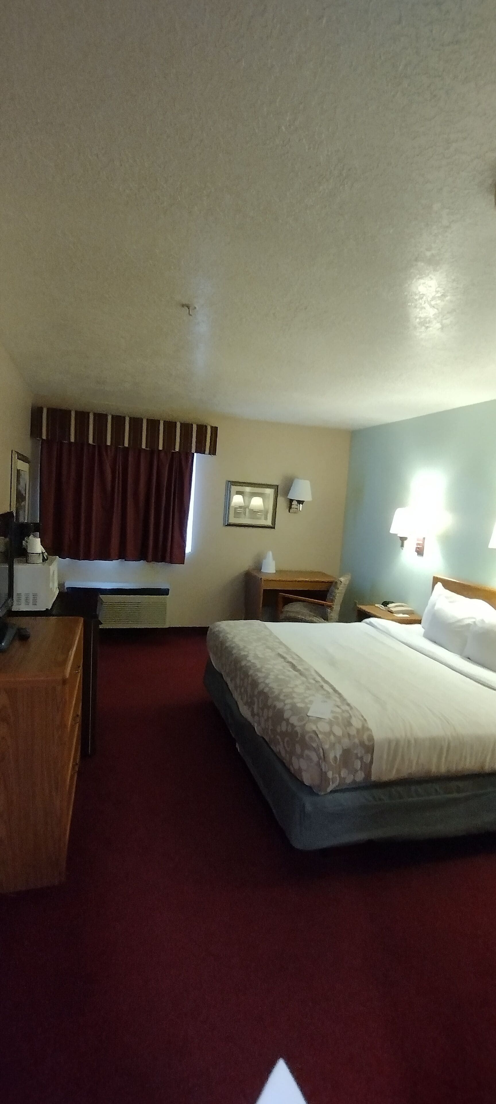 A hotel room with a bed, a desk, a chair, a lamp, and a window with curtains.