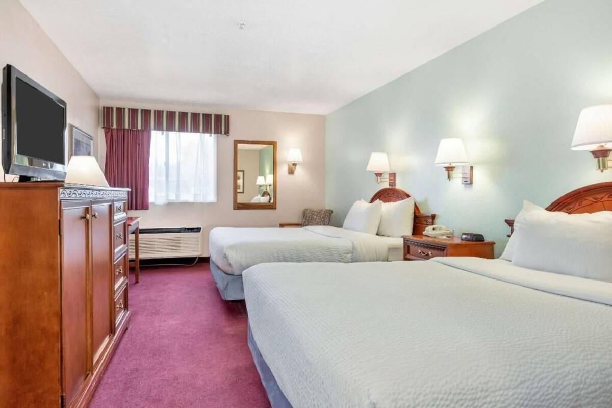 A hotel room with two beds, a television, a dresser, a mirror, and wall-mounted lamps.