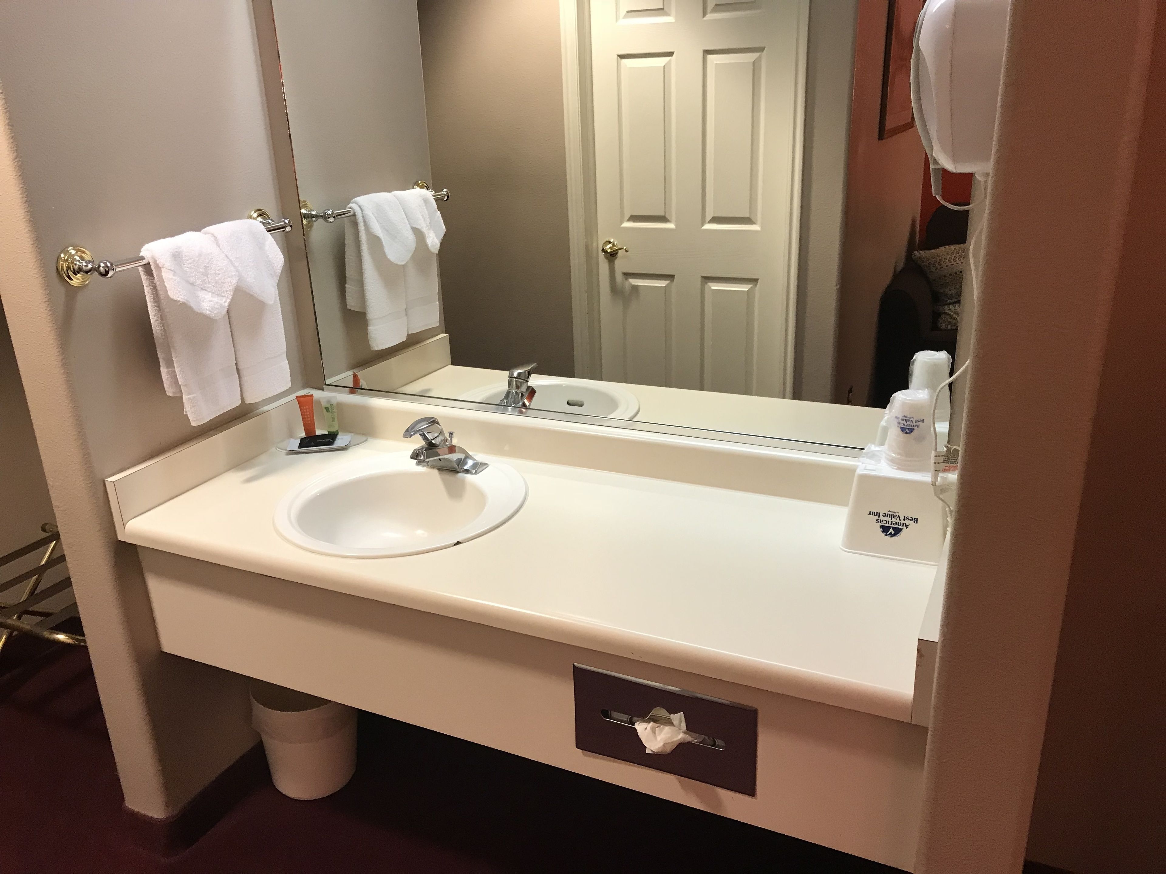 A hotel bathroom with a white sink, a mirror, a towel rack with two towels, a bottle of hand soap, and a tissue box.