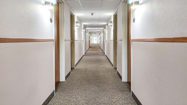 A long, narrow hotel corridor with carpeted floors and walls, flanked by doors and illuminated by wall sconces. - Americas Best Value Inn Cedar City (Cedar City)