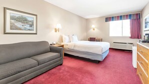 A hotel room with a bed, a sofa, a nightstand, a lamp, a dresser, and a window with curtains. - Americas Best Value Inn Cedar City (Cedar City)