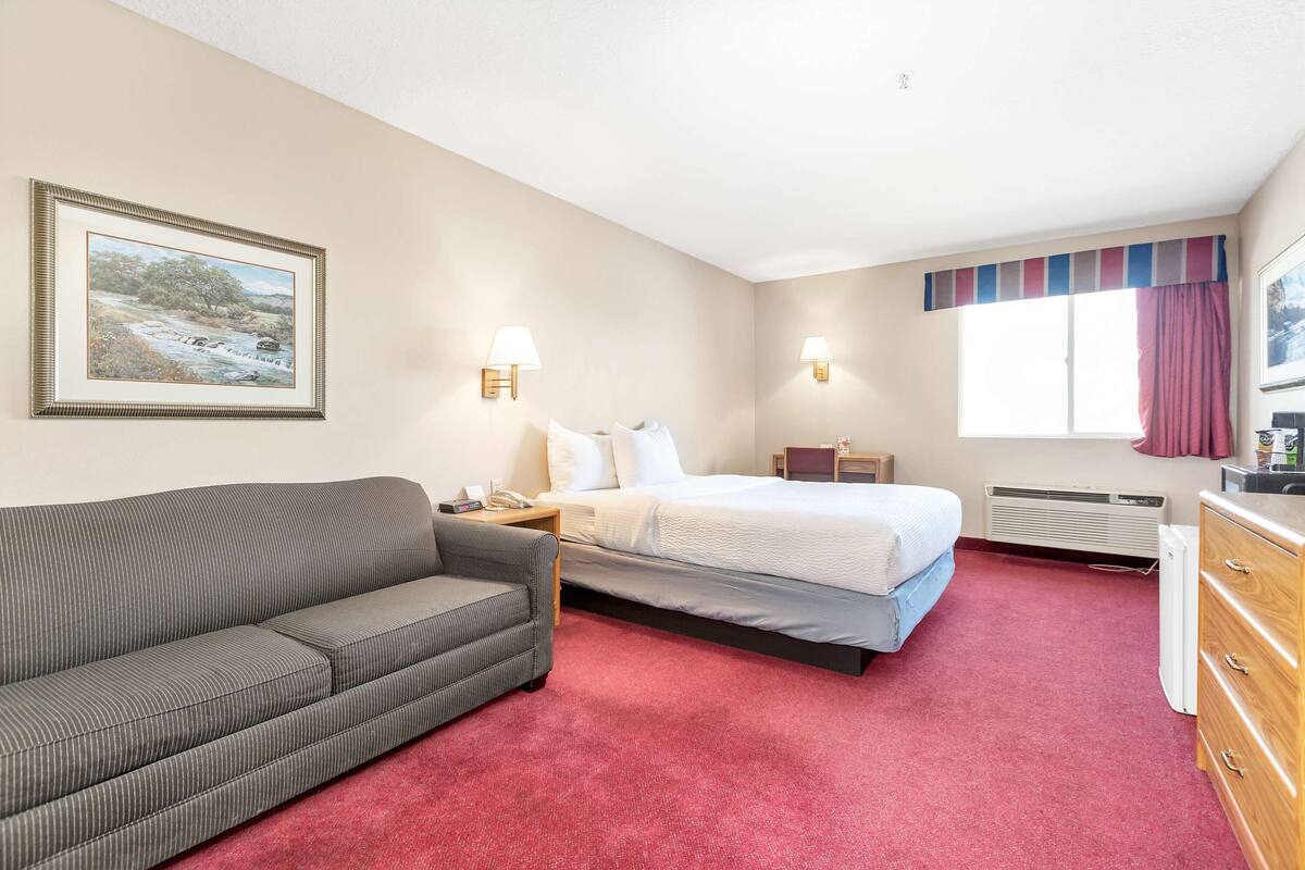 A hotel room with a bed, a sofa, a nightstand, a lamp, a dresser, and a window with curtains.