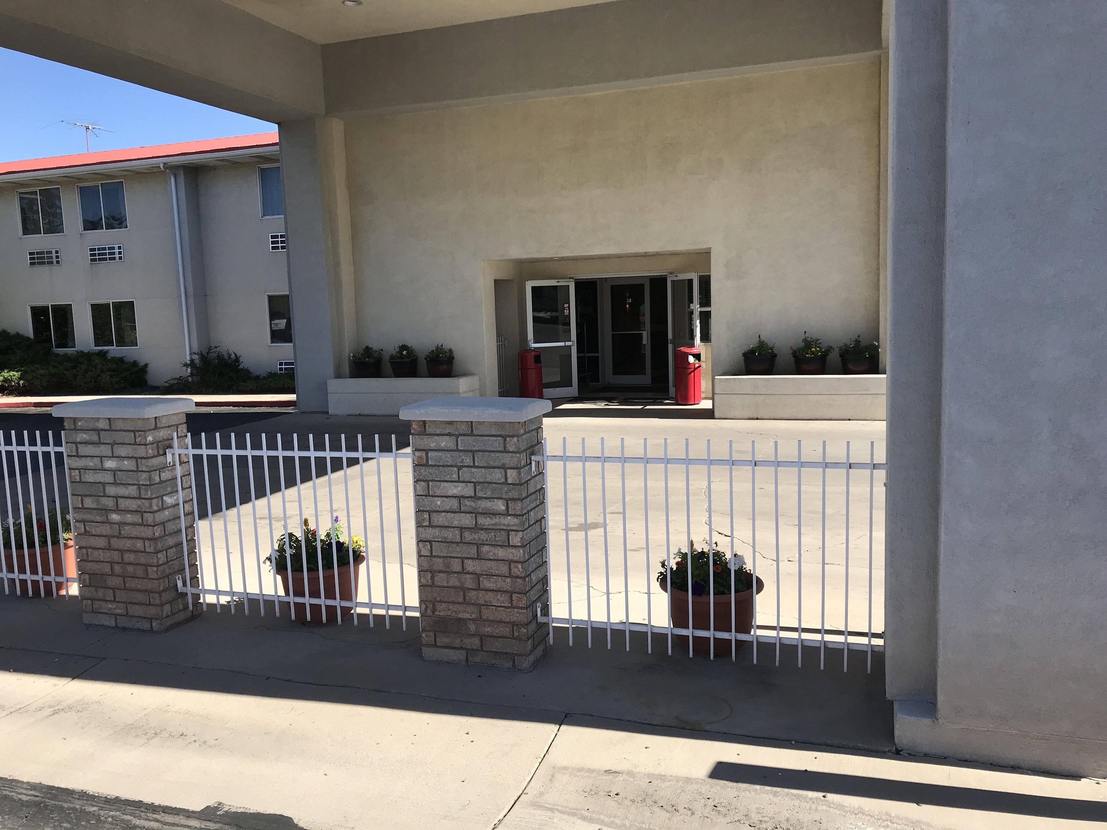 Un bâtiment doté d’une entrée fermée par un portail, de plantes en pot et d’une poubelle rouge.
