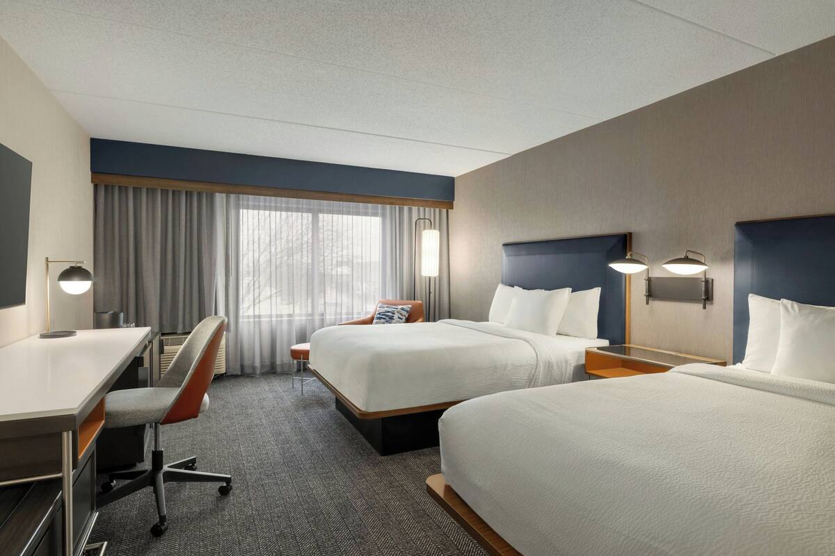 A hotel room with two beds, a desk, a chair, and a large window with curtains.