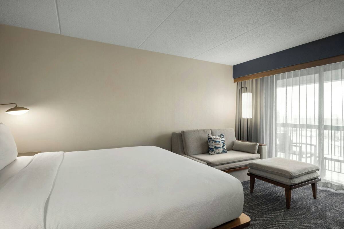 A hotel room with a large bed, a chair, a small table, and a window with curtains.