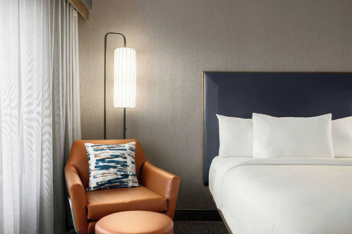 A hotel room with a bed, a chair with a pillow, a small table, and a wall-mounted lamp.