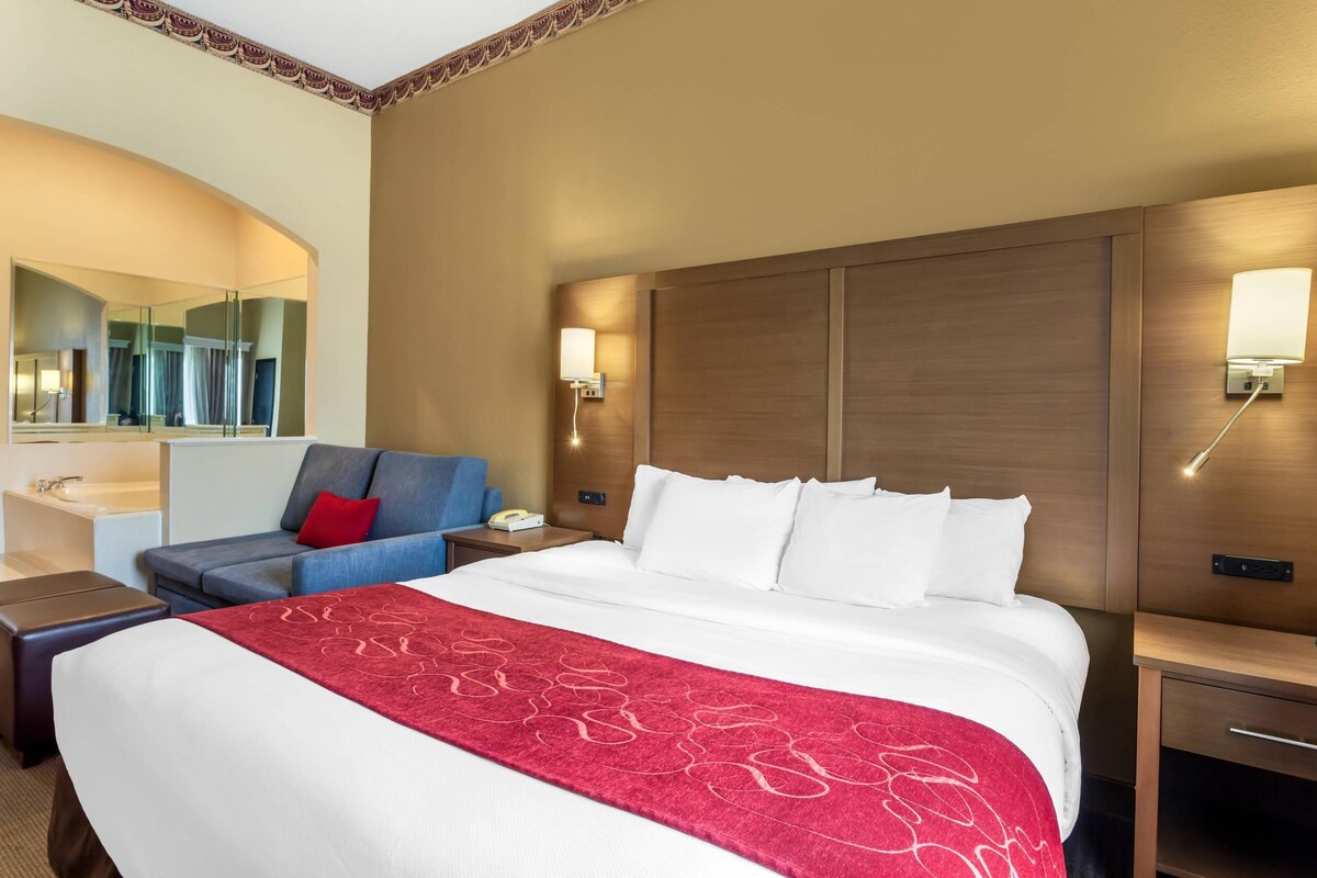 A hotel room with a large bed, a bedside table, a nightstand, a desk with a chair, a mirror, and a bathroom area.