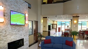 A spacious lobby with a blue sofa, a flat-screen TV mounted on a brick wall, and a dining area in the background. - Comfort Suites Bush Intercontinental Airport (Houston)