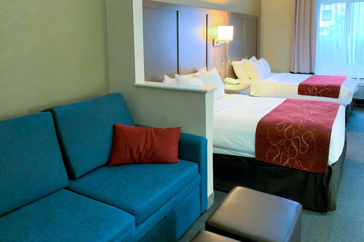 A hotel room with a blue sofa, two beds with red and white bedding, a bedside table with a lamp, and a window with curtains.