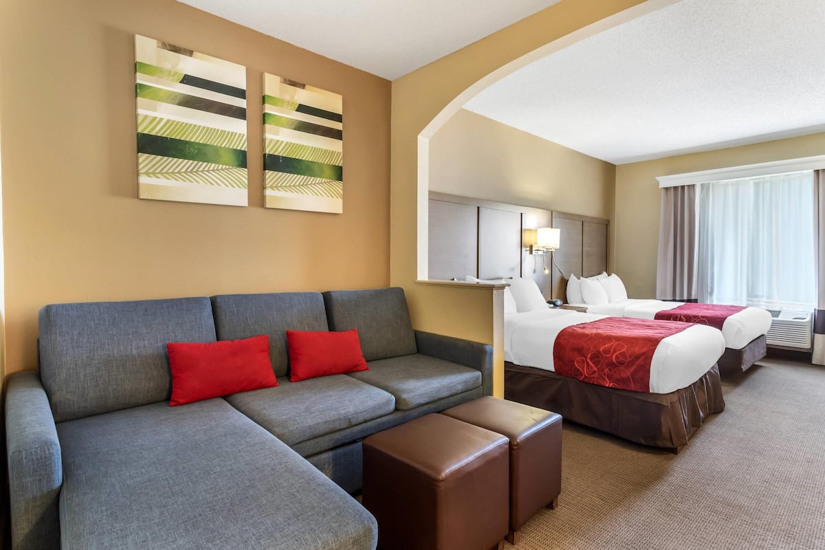 A hotel room with a grey sofa, two red pillows, a brown ottoman, a bed with a red bedspread, and a large window with curtains.