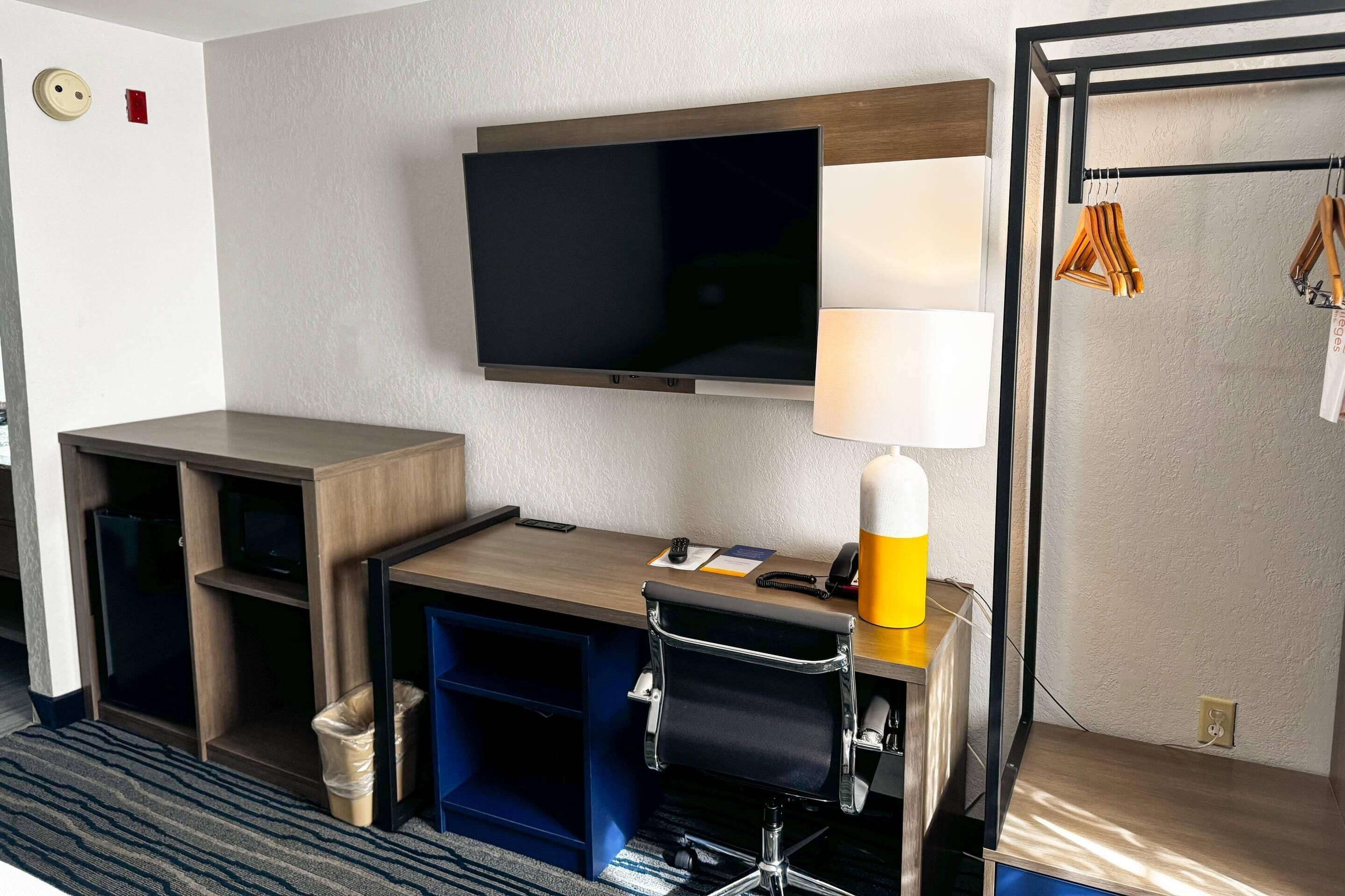 Quarto de hotel com televisão de tela plana na parede, uma mesa com abajur, uma cadeira e um cesto de lixo pequeno.