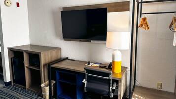 Quarto de hotel com televisão de tela plana na parede, uma mesa com abajur, uma cadeira e um cesto de lixo pequeno.