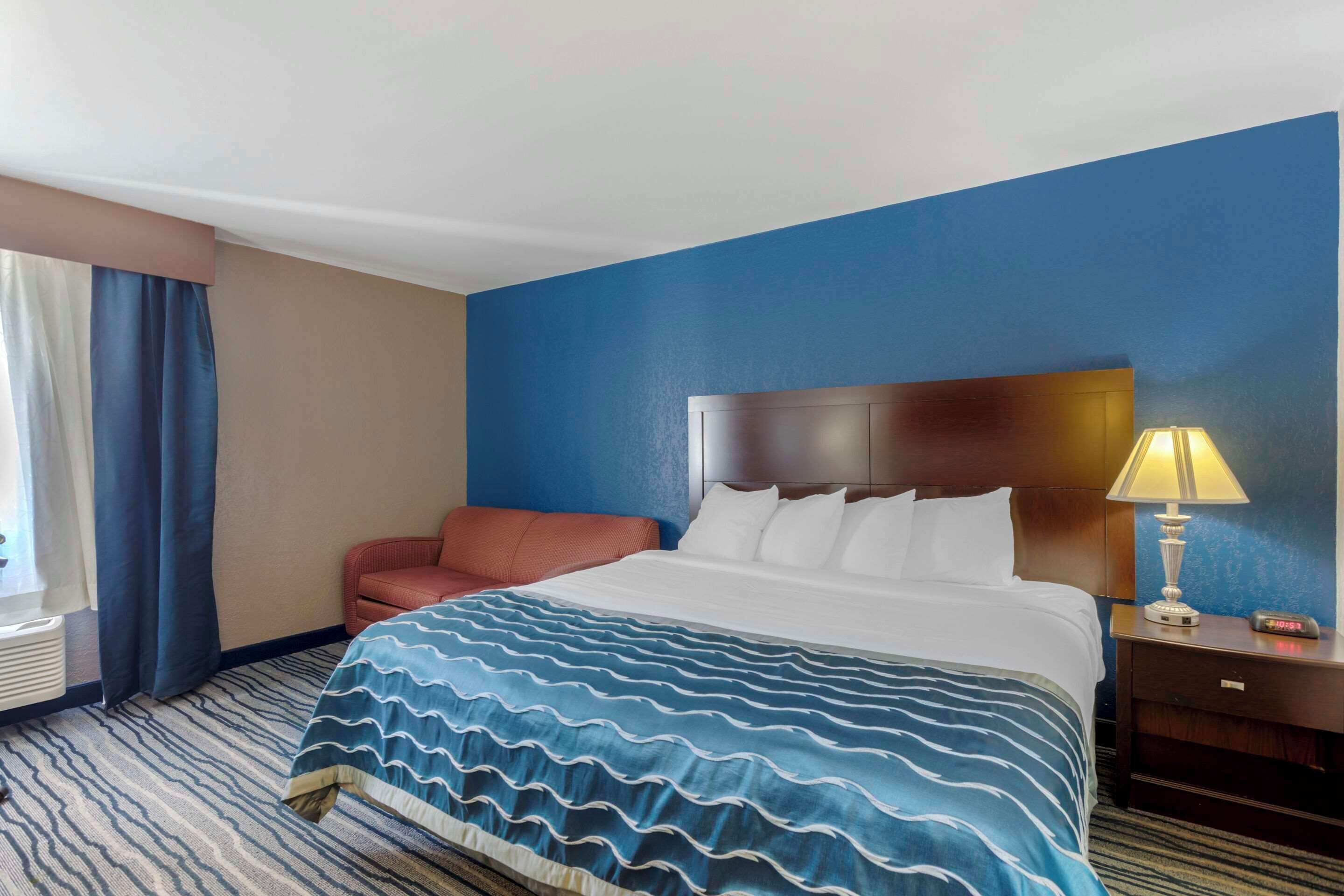 Een hotelkamer met een bed, nachtkastje met lamp, stoel en raam met gordijnen.