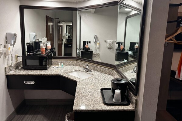 A hotel bathroom with a granite countertop, a sink, a mirror, a hair dryer, a coffee maker, and a trash bin.