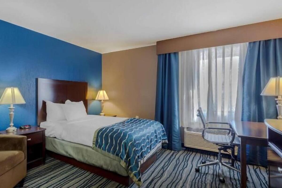 Une chambre d’hôtel avec un lit, deux lampes de chevet, un bureau, une chaise et une fenêtre avec des rideaux.