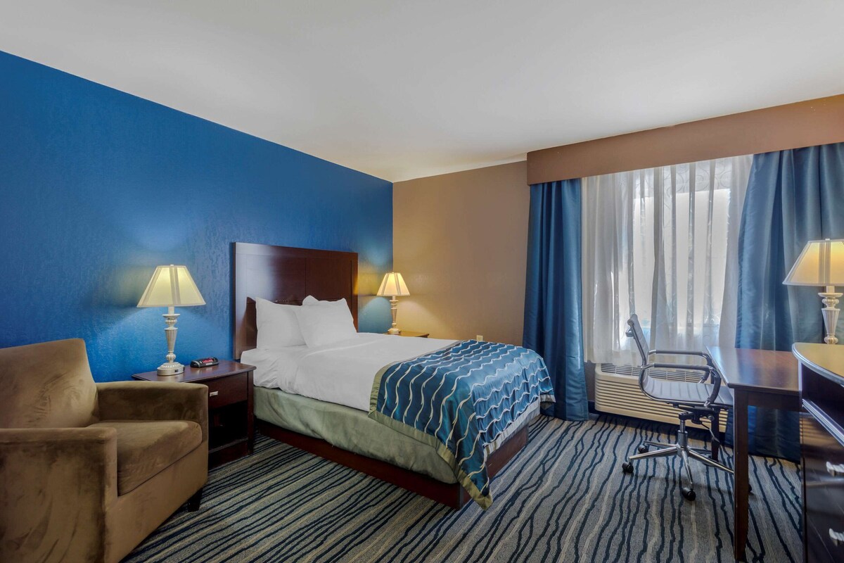 A hotel room with a bed, two bedside tables with lamps, a chair, a desk, and a large window with curtains.