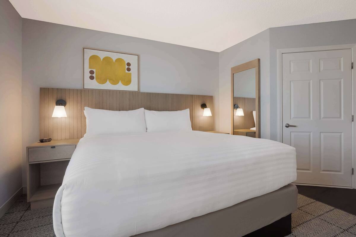 Une chambre d’hôtel avec un grand lit, deux tables de chevet avec des lampes, une tête de lit en bois, une œuvre d’art accrochée au mur et une porte.