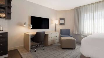 A hotel room with a bed, a desk with a chair, a television, a lamp, and a small ottoman.
