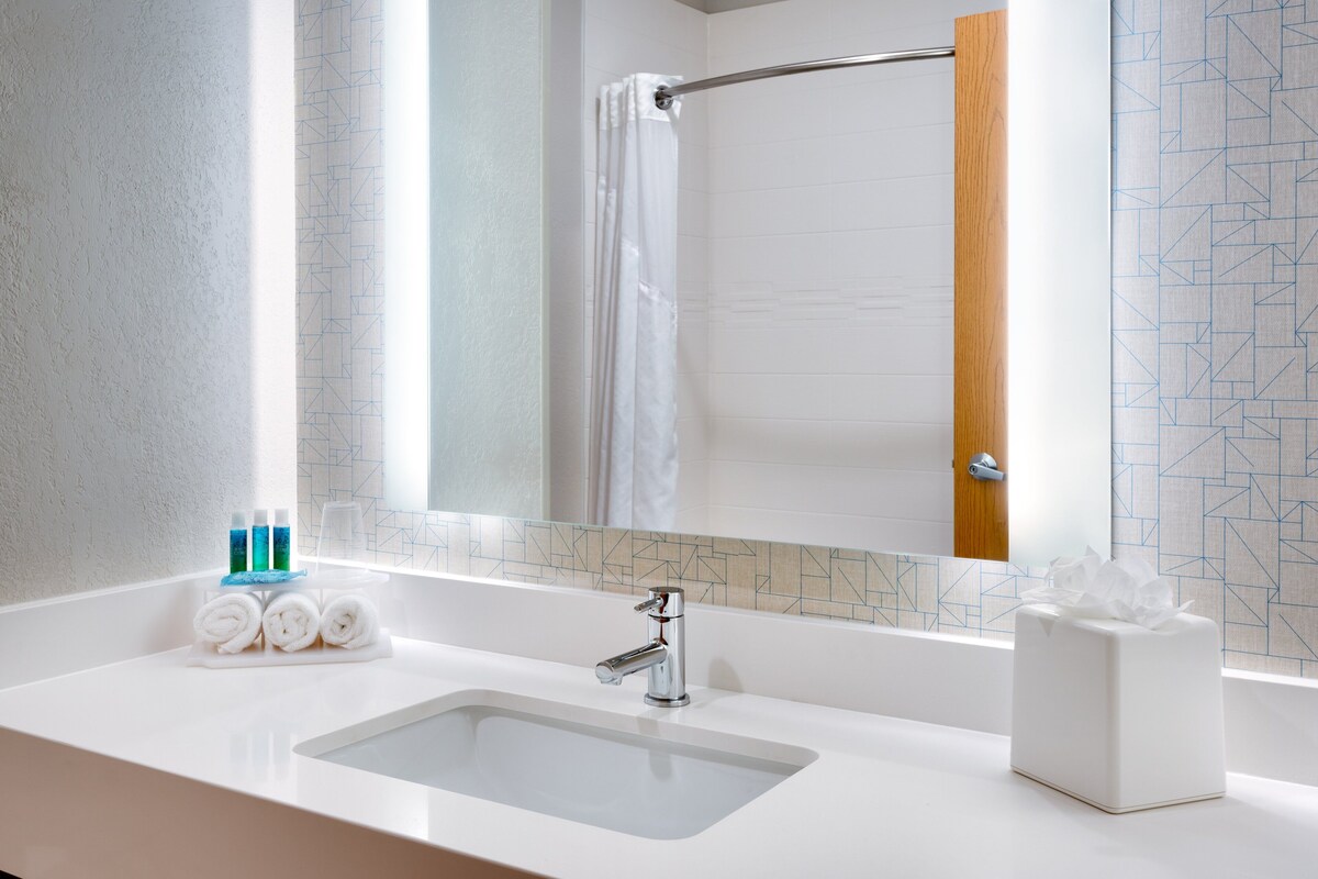 A hotel bathroom with a white sink, a mirror, a shower curtain, and toiletries on the counter.