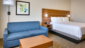 Quarto de hotel com cama, sofá azul, mesa de centro de madeira, mesa de cabeceira com telefone e uma obra de arte abstrata emoldurada na parede.