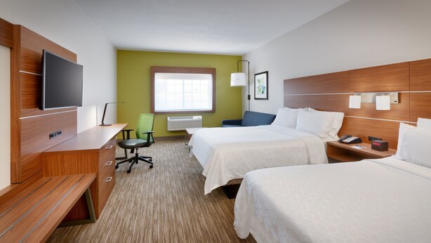 Quarto de hotel com duas camas, uma escrivaninha, televisão de tela plana, uma cadeira verde e uma janela com persianas.