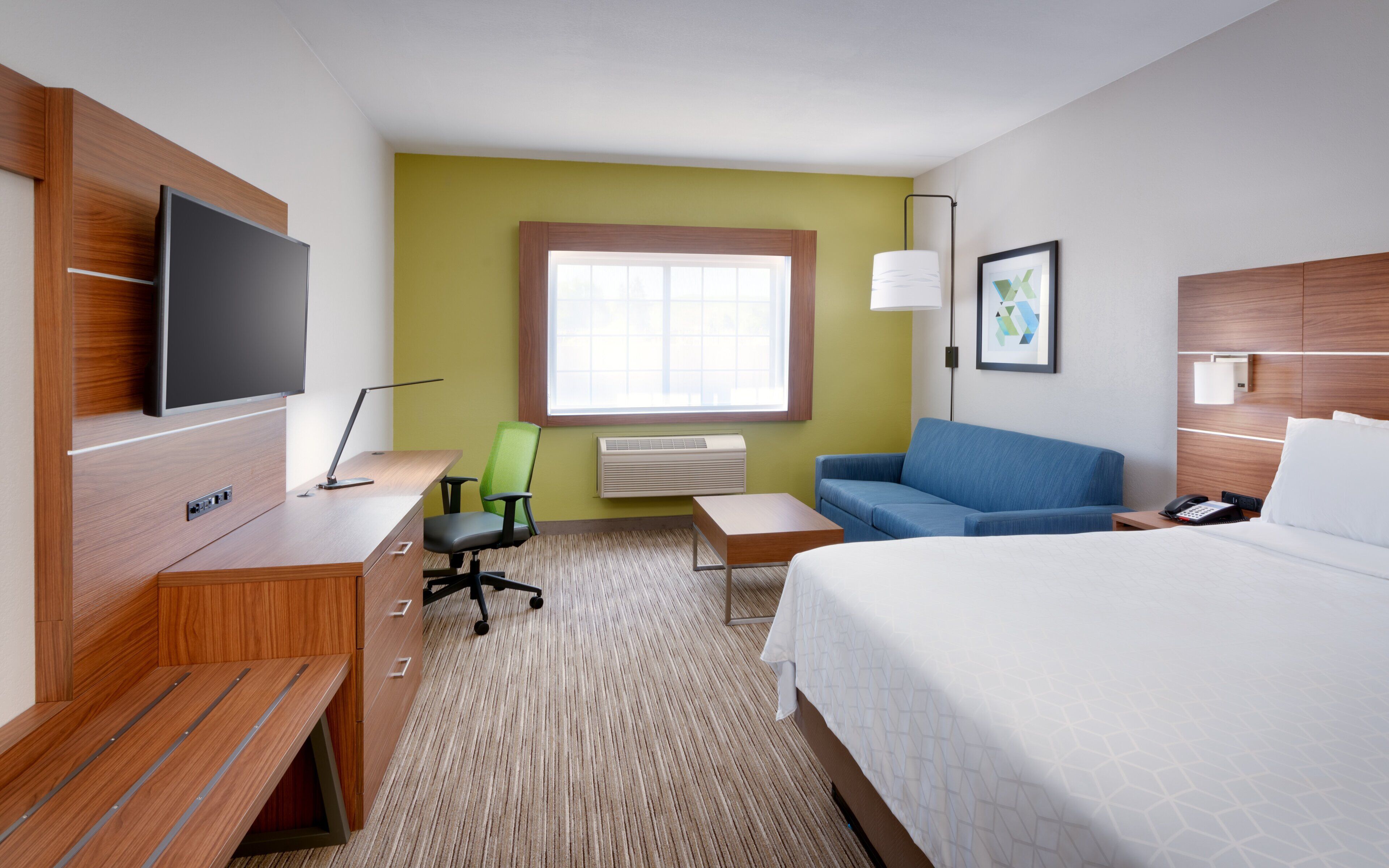 A hotel room with a bed, a desk with a chair, a sofa, a TV, a window, and a lamp.
