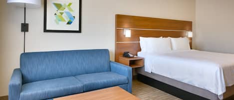 Habitación de hotel con una cama, un sofá azul, una mesita de noche de madera, una lámpara y una obra de arte abstracta enmarcada.