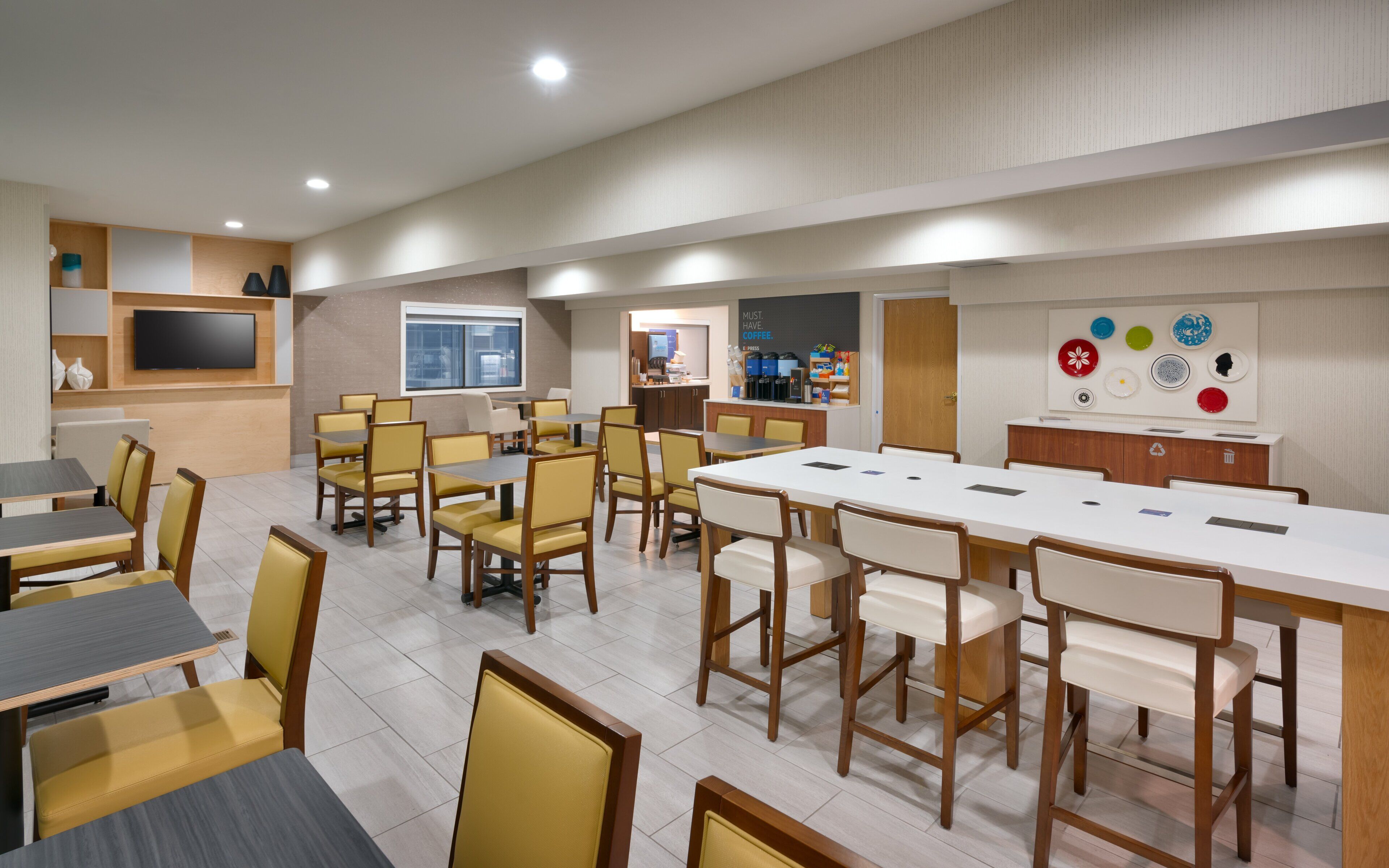 Uma sala de jantar moderna com várias mesas e cadeiras, uma TV de tela plana na parede e um espaço com cafeteira e diversos itens.