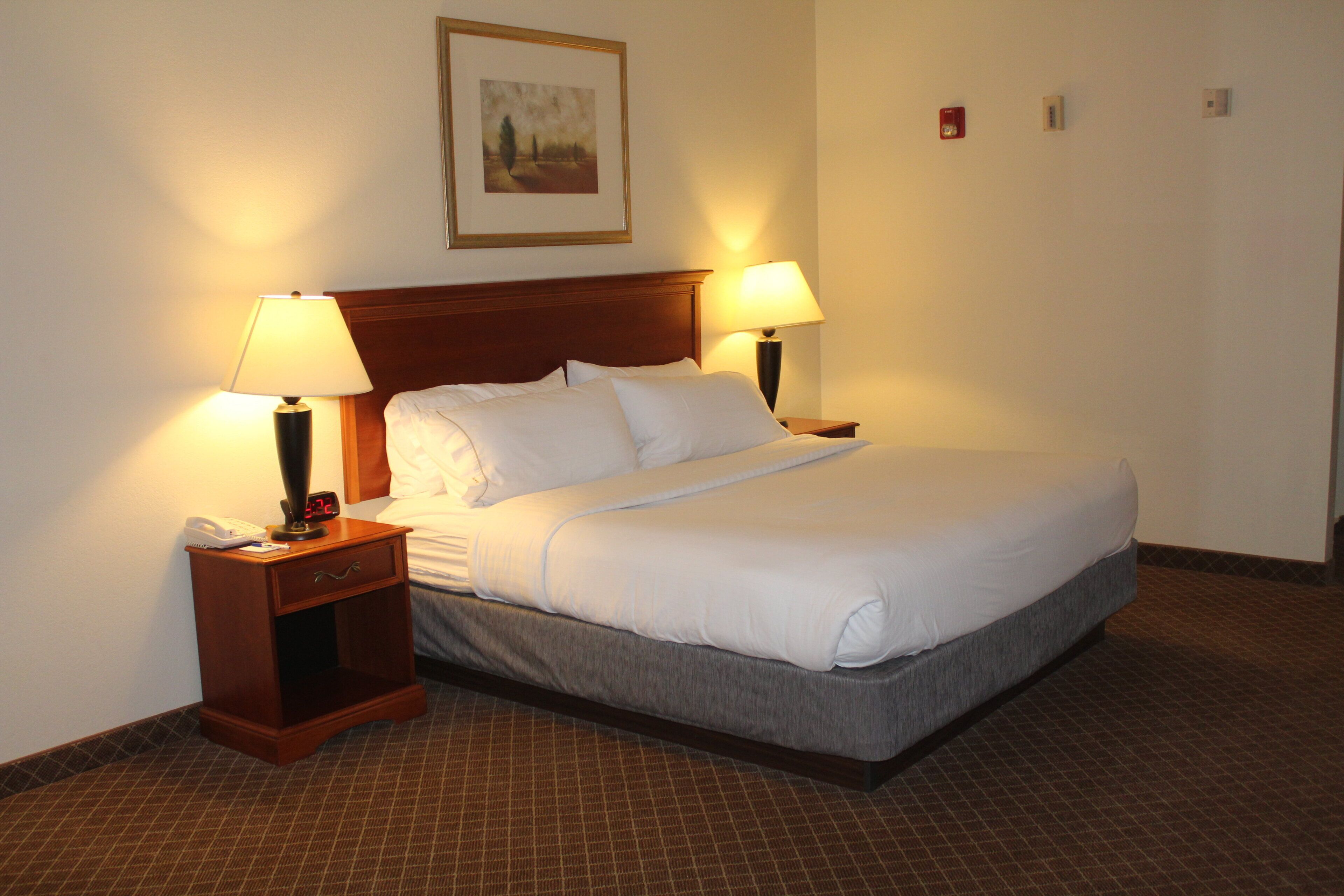 A neatly made bed with white linens, two bedside lamps, a wooden nightstand with a phone and a clock, and a framed picture on the wall.