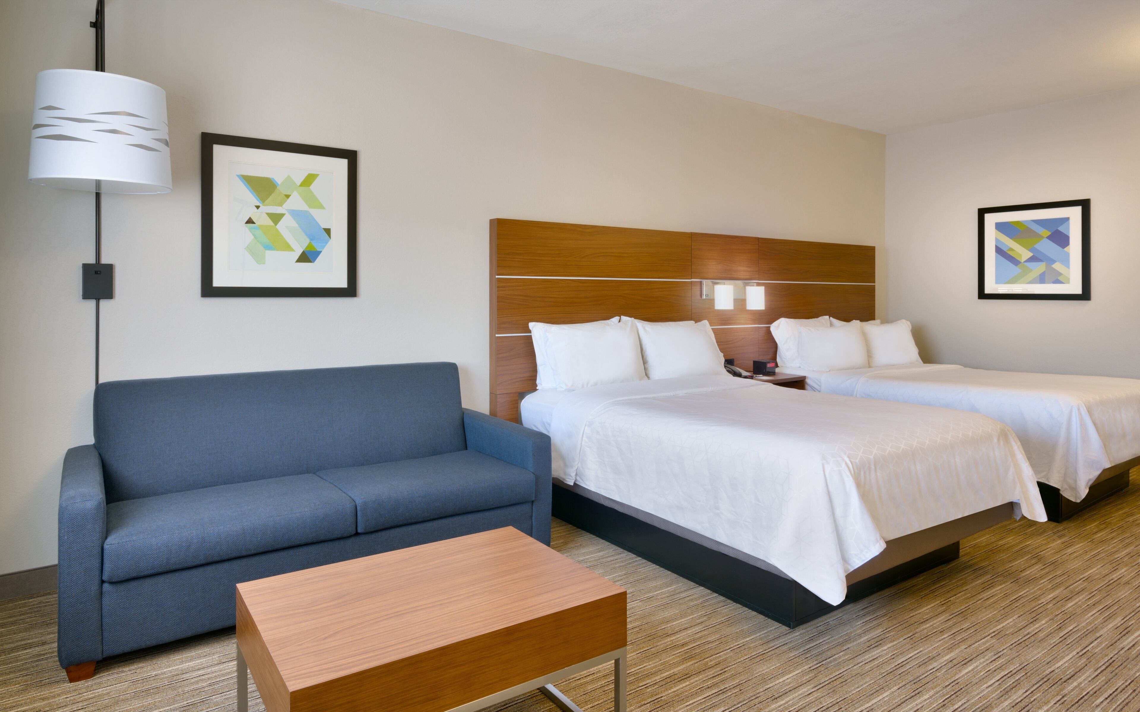 Habitación de hotel con un sofá azul, una mesa de centro de madera, dos camas con ropa blanca, un cabecero de madera y dos obras de arte abstracto enmarcadas en las paredes.