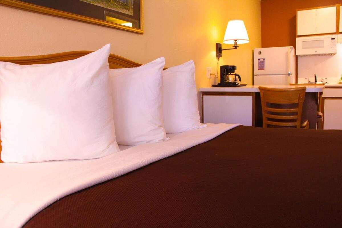 A hotel room with a bed, pillows, a desk, a chair, a lamp, a refrigerator, and a microwave.