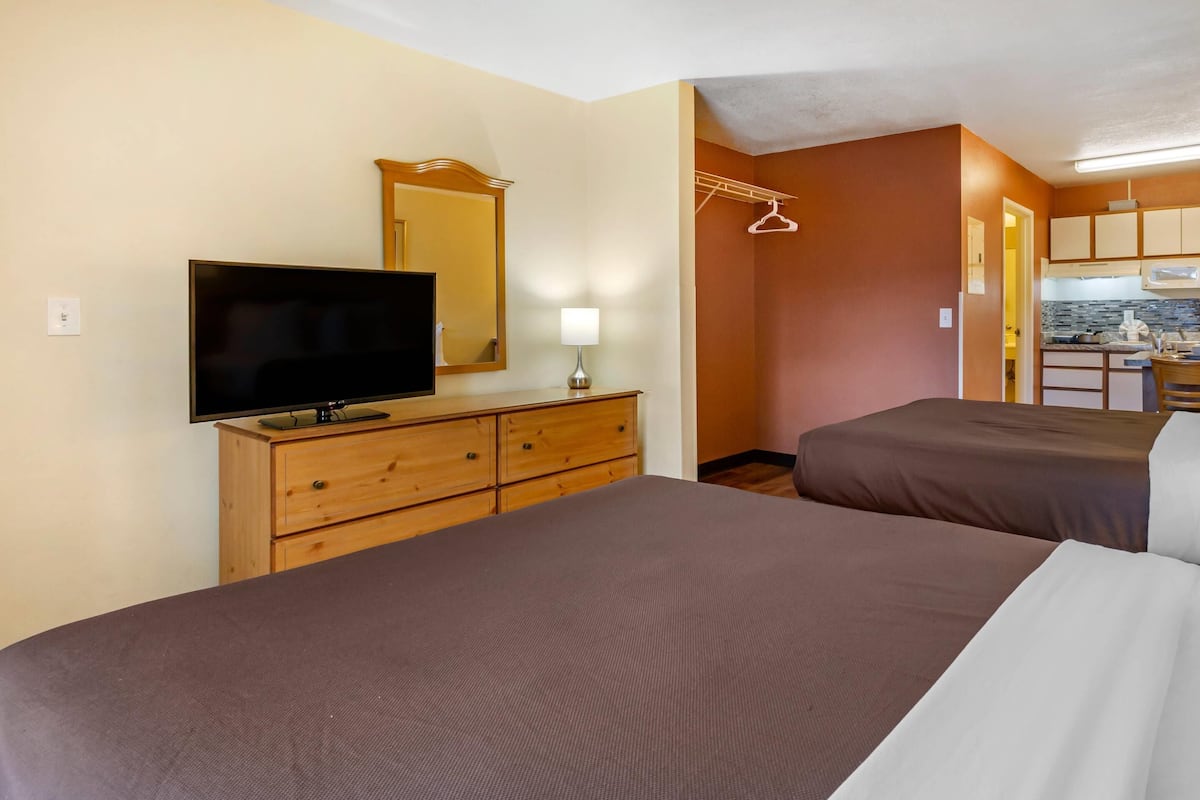 A hotel room with two beds, a TV on a wooden dresser, a mirror, and a kitchenette in the background.