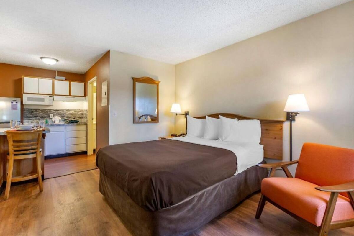 A hotel room with a bed, a desk, a chair, a lamp, and a kitchenette.