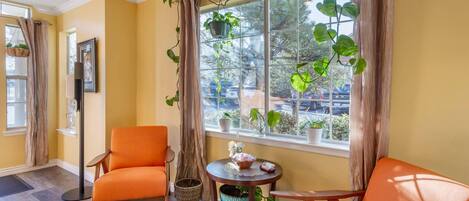 A room with a yellow wall, a large window with curtains, a chair, a small table with a vase, and a potted plant.