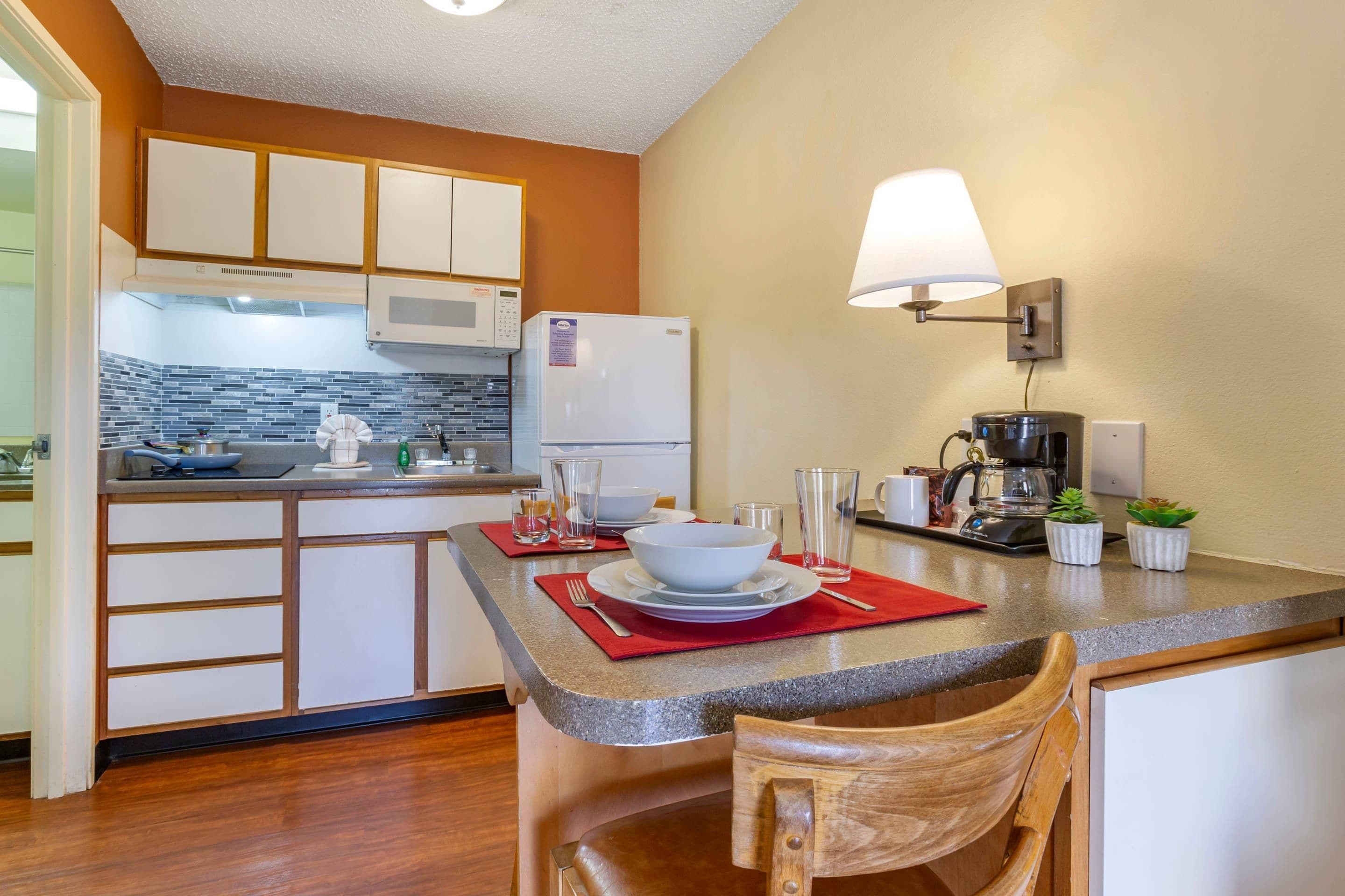 A compact kitchen with a microwave, refrigerator, and coffee maker. There is a dining area with a chair and a table set for two.