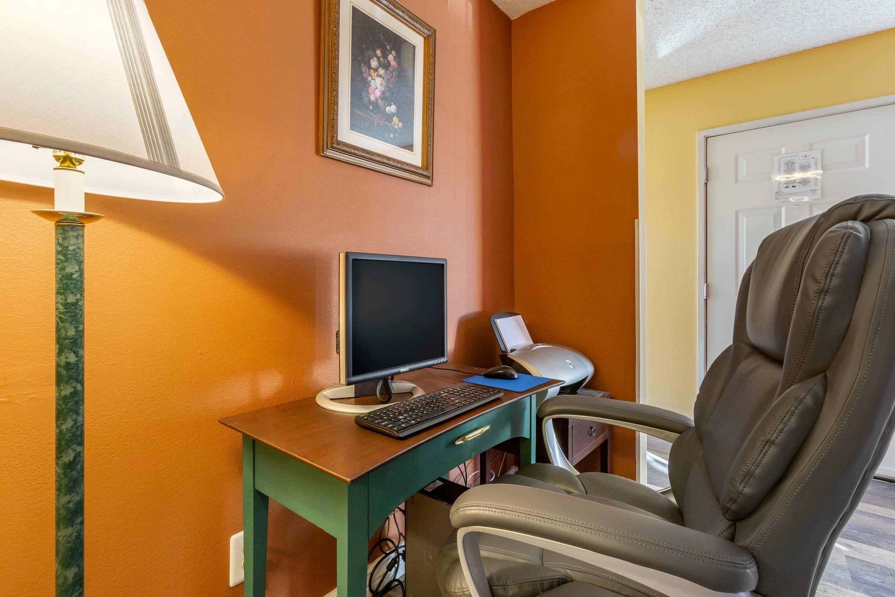 A room with a desk, computer, chair, lamp, and a framed picture on the wall.