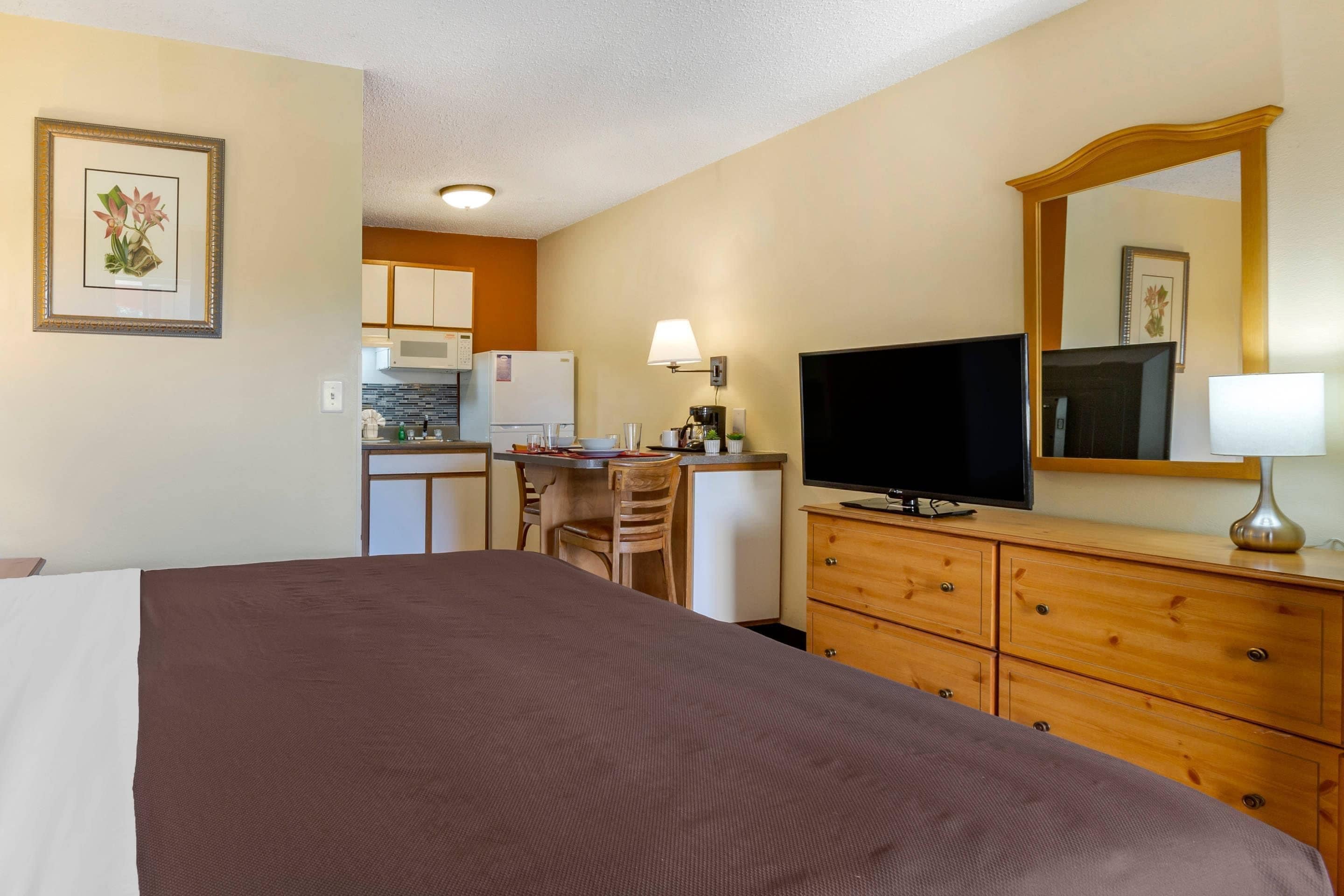 A hotel room with a bed, a desk, a television, a dresser, and a kitchenette in the background.
