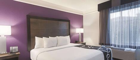 A hotel room with a large bed, two bedside tables, a nightstand, a window with curtains, and an air conditioning unit.