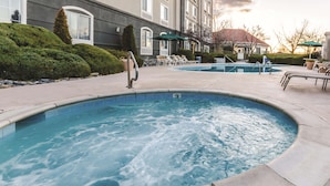 Un jacuzzi rodeado de sillas de descanso y una piscina, con un edificio al fondo.