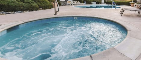 A hot tub surrounded by lounge chairs and a pool, with a building in the background.