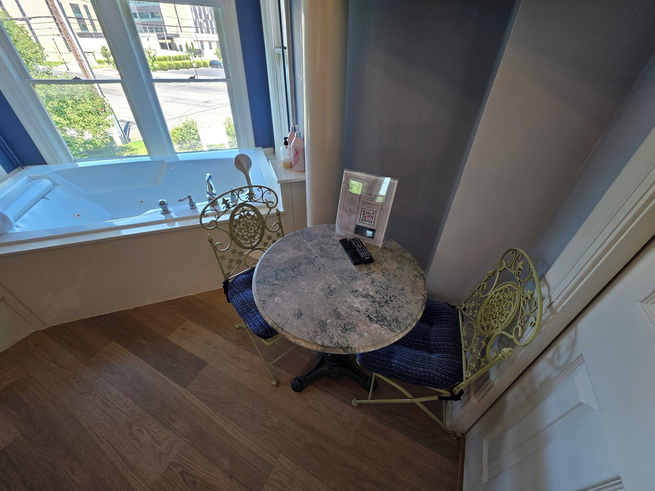 Een hoekkamer met een ronde marmeren tafel, twee stoelen, een raam met uitzicht op de straat en een kleine tafel met een afstandsbediening en een QR-code.